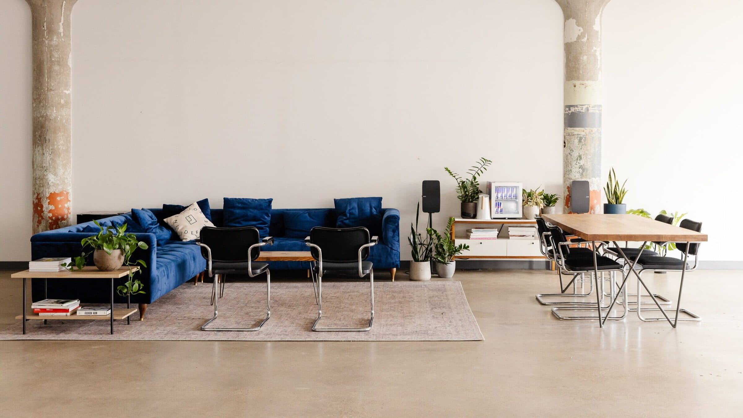 The studio lounge: a blue velvet sofa on a rug, black cantilever chairs, a long wooden table, plants, and Sonos speakers
